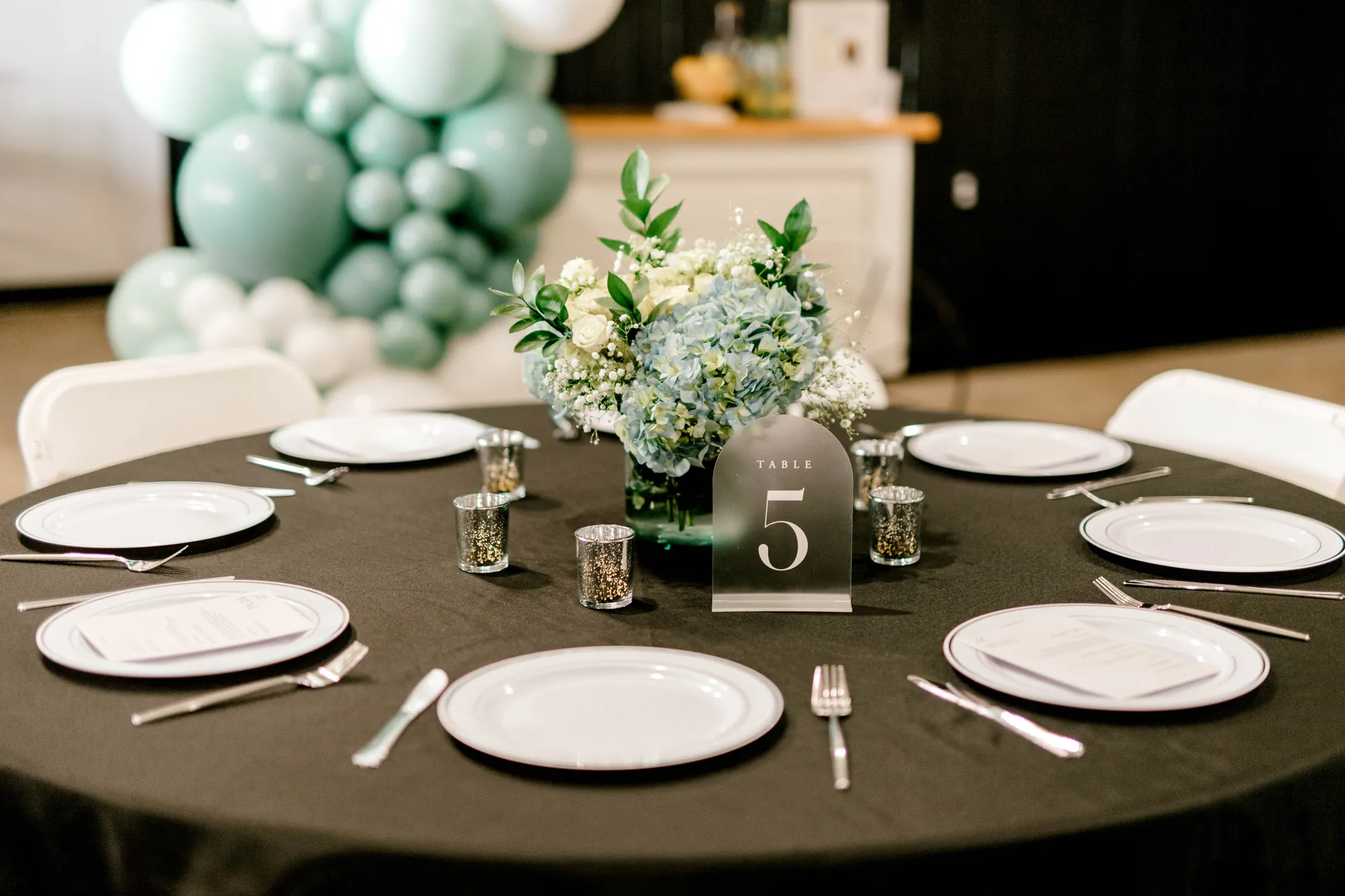 A round table with black linen and a sage balloon garland in the hall